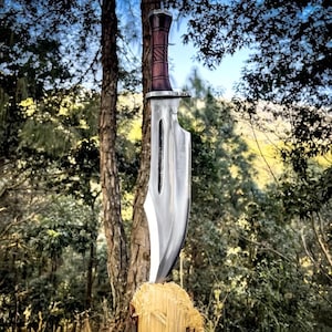 Può includere: Un grande coltello in acciaio lucido con un manico in legno scuro, conficcato in un ceppo d'albero. La lama ha una forma distintiva con un ritaglio vicino al manico. Il manico è avvolto con un materiale marrone scuro strutturato. Lo sfondo presenta un ambiente forestale.