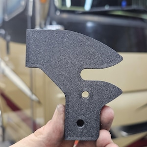 May include: A black, textured, 3D-printed bracket with a unique shape. It features a curved top, a central cutout, and two circular holes. The bracket is held up against a blurred background.