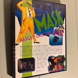 Puede incluir: Póster enmarcado del videojuego "The Mask" con Madcap Cool Cat. El póster presenta un personaje con traje amarillo, el título del juego y un perro. El texto incluye "Action-game fans will shout 'Smokin'!"