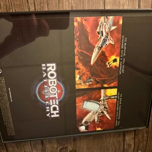 May include: Framed artwork featuring the "Robotech Battlecry" logo and images of futuristic fighter jets in a fiery, action-packed scene. The artwork includes text that reads "To survive, you must change into a warrior" and "To save humankind, you must change into a hero."