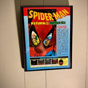 May include: Framed Spider-Man poster featuring the text "Return of the Sinister Six." The artwork showcases Spider-Man's mask with villains reflected in the eyes. The poster has a red, blue, and yellow color scheme, with game instructions at the bottom.