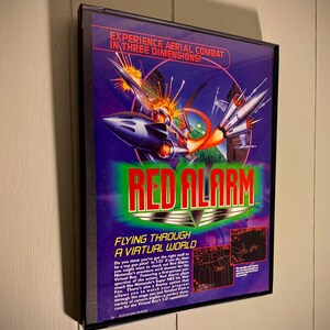 Könnte beinhalten: Gerahmte Red Alarm Videospielbox. Das Artwork zeigt ein futuristisches Raumschiff, das Raketen abfeuert, mit dem Text "EXPERIENCE AERIAL COMBAT IN THREE DIMENSIONS!" und "RED ALARM". Das Spiel ist für den Nintendo Virtual Boy.