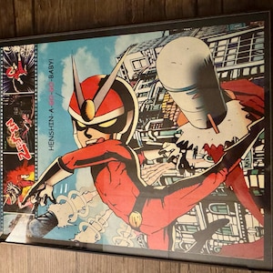 May include: Framed comic book art featuring a superhero in a red suit and helmet, with the text "HENSHIN-A-GO-GO-BABY!". The artwork has a retro comic book style with a blue sky background and a cityscape.