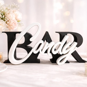 Puede incluir: Letrero de madera en blanco y negro con la palabra BAR y la palabra "Candy" en cursiva. Velas, flores y macarons en el fondo. El letrero es un elemento decorativo para una barra de dulces o un evento.
