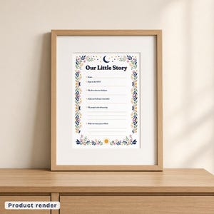The Our Little Story page in a light wood frame with a wide white mat, standing on a light wood dresser against a cream wall. The page has a navy title, a crescent moon and stars at the top, a sun at the bottom, a border of leaves and flowers, and six labeled spaces with blank lines.