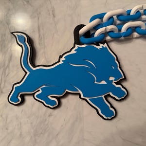 May include: A three-dimensional wall hanging featuring a blue and white lion logo in a leaping pose, outlined in black. The lion is attached to a decorative chain made of interlocking white and blue plastic links, creating a layered effect.