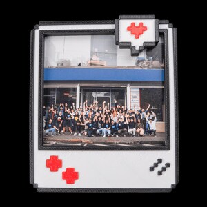 May include: A white and black framed photo featuring a group of people, with red pixelated heart and cross symbols. The frame has a retro video game controller design. The photo shows a building exterior with a blue awning.