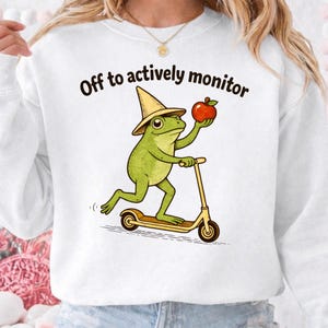 May include: White sweatshirt with a graphic of a green frog wearing a hat and riding a scooter while holding a red apple. The text "Off to actively monitor" is above the graphic.
