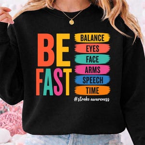 Peut inclure: Sweat-shirt noir avec les mots "BE FAST" en orange, jaune et turquoise. En dessous, les mots "BALANCE, EYES, FACE, ARMS, SPEECH, TIME" sont listés dans des blocs colorés. Le texte "#stroke awareness" est en bas.