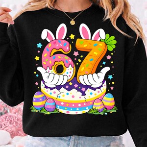 Pode incluir: Camisola preta com um gráfico colorido com tema de Páscoa. O design apresenta o número 67 feito de um donut e uma cenoura, orelhas de coelho e um ovo com listras coloridas. Ovos de Páscoa e estrelas também estão incluídos.