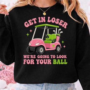 Pode incluir: Camisola preta com um desenho de carrinho de golfe rosa e verde. O texto "GET IN LOSER WE'RE GOING TO LOOK FOR YOUR BALL" está em rosa e verde. O design inclui estrelas.