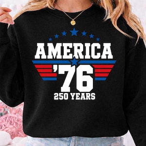 May include: A black long-sleeve sweatshirt featuring a graphic print with the text AMERICA '76 250 YEARS in white, accented by a blue star and horizontal red and blue stripes, with a small arc of blue stars positioned above the word AMERICA.