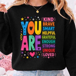 Op de afbeelding: Zwarte sweatshirt met de woorden "YOU ARE" in kleurrijke blokletters. De woorden "KIND, BRAVE, SMART, HELPFUL, GRATEFUL, ENOUGH, STRONG, UNIQUE, LOVED" zijn verticaal gestapeld in regenboogkleuren.