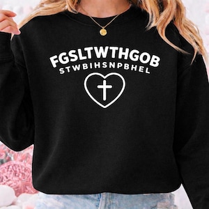 Könnte beinhalten: Schwarzes Sweatshirt mit weißem Text und einer herzförmigen Umrandung mit einem Kreuz. Der Text oben lautet "FGSLTWTHGOB" und der Text darunter lautet "STWBIHSNPBHEL". Eine goldene Halskette ist sichtbar.