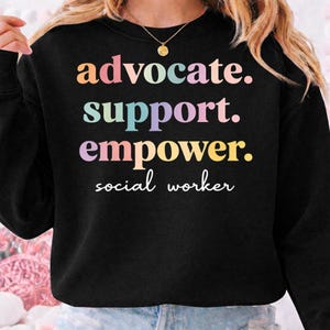 May include: Black sweatshirt with the words "advocate. support. empower. social worker" in colorful, pastel lettering. The design is centered on the chest. A gold necklace is visible at the neckline.