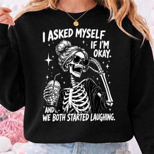 May include: Black sweatshirt with a graphic of a skeleton wearing sunglasses and holding a drink. The text reads "I ASKED MYSELF IF I'M OKAY. AND WE BOTH STARTED LAUGHING." The design includes stars and a bun hairstyle.