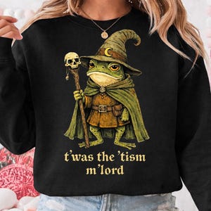 Op de afbeelding: Zwart sweatshirt met een fantasie-thema. Het toont een kikker in een tovenaarsoutfit, compleet met hoed, cape en staf met een schedel. De tekst "t'was the 'tism m'lord" is in goud gedrukt. De afbeelding staat in het midden op de voorkant.