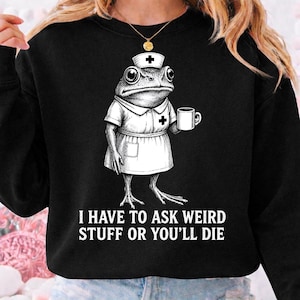 May include: Black sweatshirt featuring a cartoon frog dressed as a nurse, holding a mug. The frog wears a nurse's cap and uniform with a cross emblem. The text "I HAVE TO ASK WEIRD STUFF OR YOU'LL DIE" is printed below the frog.