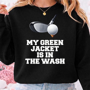 May include: Black sweatshirt with a golf club and golf ball graphic. The text "MY GREEN JACKET IS IN THE WASH" is printed in white. A gold pendant necklace is visible.