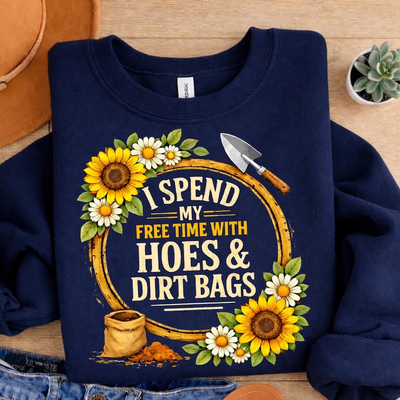 May include: Navy blue sweatshirt with the text "I Spend My Free Time With Hoes & Dirt Bags." The design includes sunflowers, daisies, and a small shovel. A gardening-themed apparel item.