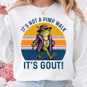 Op de afbeelding: Een wit sweatshirt met ronde hals en een afbeelding van een groene kikker in een paarse bontjas, een luipaardhoed en een gouden dollarteken-ketting. De kikker houdt een wandelstok vast. De tekst luidt IT'S NOT A PIMP WALK IT'S GOUT! op een cirkel.