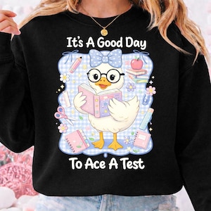 May include: Black sweatshirt with a cartoon duck wearing glasses and a bow, reading a book. The text "It's A Good Day To Ace A Test" is displayed above and below the duck, surrounded by school-themed illustrations.