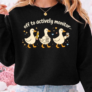 May include: Black sweatshirt with a whimsical design featuring three cartoon ducks. The ducks are walking and holding a pencil, a shirt with the text "YOU GOT THIS", and a coffee cup. The text "off to actively monitor..." is above the ducks.