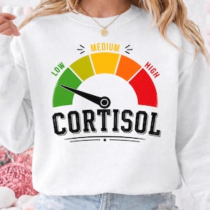 May include: White sweatshirt featuring a gauge design with the word "CORTISOL" in bold, black letters. The gauge displays "LOW," "MEDIUM," and "HIGH" in green, yellow, and red, respectively. A black needle points towards the "LOW" end.