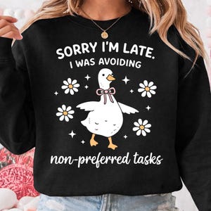 May include: Black sweatshirt with a white goose graphic wearing a pink bow tie. The text reads "SORRY I'M LATE, I WAS AVOIDING non-preferred tasks" with daisy and star accents. A casual, humorous design.