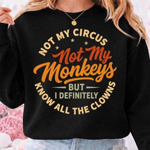 以下が含まれることがあります： 黒いスウェットシャツ。オレンジと黄色の文字で「NOT MY CIRCUS, NOT MY MONKEYS, BUT I DEFINITELY KNOW ALL THE CLOWNS」と書かれています。デザインは胸の中央に配置されています。