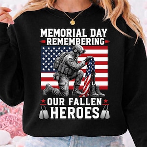 May include: Black sweatshirt with a graphic commemorating Memorial Day. The design shows a kneeling soldier, the American flag, and the text "MEMORIAL DAY REMEMBERING OUR FALLEN HEROES" in white. Includes dog tags and red stars.