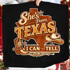K&ouml;nnte beinhalten: Schwarzes T-Shirt mit einer braunen Grafik, die den Bundesstaat Texas, einen Cowboyhut und einen Cowboy-Stiefel zeigt. Der Text lautet "She's From Texas I Can Tell" im Vintage-Stil.