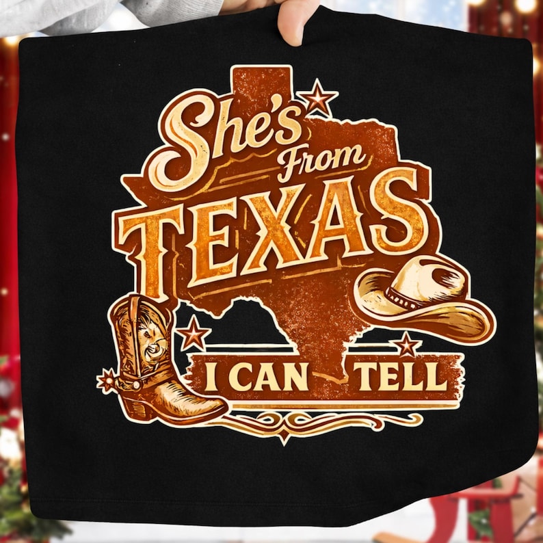 K&ouml;nnte beinhalten: Schwarzes T-Shirt mit einer braunen Grafik, die den Bundesstaat Texas, einen Cowboyhut und einen Cowboy-Stiefel zeigt. Der Text lautet "She's From Texas I Can Tell" im Vintage-Stil.