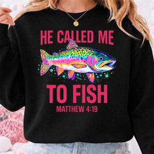 Può includere: Felpa nera con la scritta rosa "HE CALLED ME TO FISH" e la citazione biblica Matteo 4:19. Un motivo di trota iridea multicolore adorna il davanti, con colori vivaci.