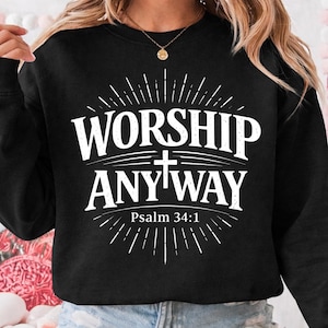 May include: Black sweatshirt with the words "WORSHIP ANYWAY" in white, with a cross and the text "Psalm 34:1". The design is surrounded by radiating lines.