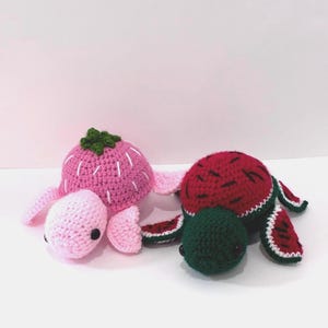 May include: Two crocheted turtle plushies. One is pink with white accents and a green top, resembling a strawberry. The other is red with green and white accents, resembling a watermelon. Both have black eyes and are on a white surface.