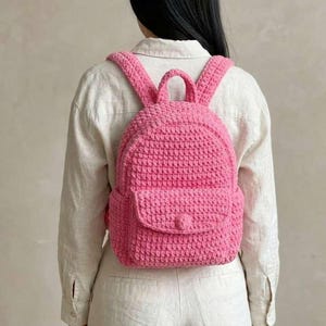 Peut inclure: Un sac à dos rose en crochet avec des bretelles réglables et une poignée supérieure. Le sac à dos a une poche avant avec une fermeture à bouton. Le sac est fait d'un fil doux et texturé.