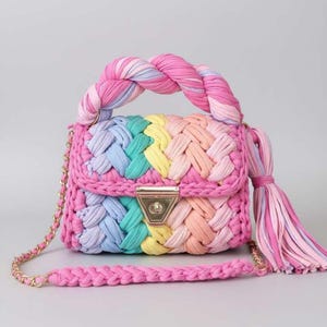 May include: A colorful, handmade handbag with a braided design. The bag features a rainbow of colors, including pink, blue, yellow, and orange. It has a gold chain strap and a matching braided handle. A pink tassel hangs from the side.