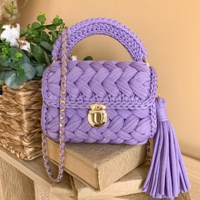 May include: A lavender crochet handbag with a braided design, a gold chain strap, and a matching tassel. The bag features a gold clasp and a braided handle. The bag is sitting on a book.