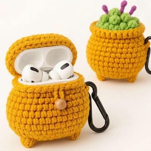 May include: Two yellow crochet cases shaped like cauldrons, one open with white earbuds inside. The other cauldron is closed and has green and purple crochet details on top. Both have black carabiner clips.