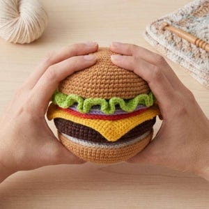 Könnte beinhalten: Ein gehäkelter Hamburger mit einem braunen Brötchen, grünem Salat, gelbem Käse und einem braunen Patty. Der Burger wird von zwei Händen gehalten. Der Burger ist handgefertigt.