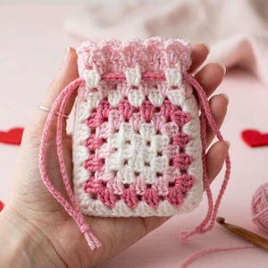 May include: A small, handmade crochet drawstring bag in white, pink, and rose. The bag features a granny square design with a ruffled top and pink drawstring. The bag is held in a hand, with a pink background and heart-shaped decorations.