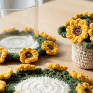 May include: Handmade crochet sunflower coasters and a small vase with a sunflower arrangement. The coasters have a green and white base with yellow sunflower accents. The vase is beige and holds several yellow sunflowers.