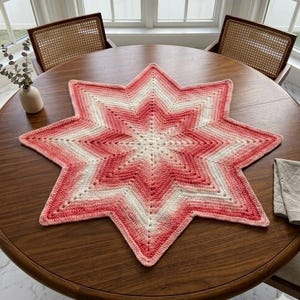 May include: A handmade, star-shaped crochet doily in shades of red, white, and pink. The doily is placed on a round wooden table. The star has eight points and a detailed openwork design. The table is surrounded by two chairs with woven backs.