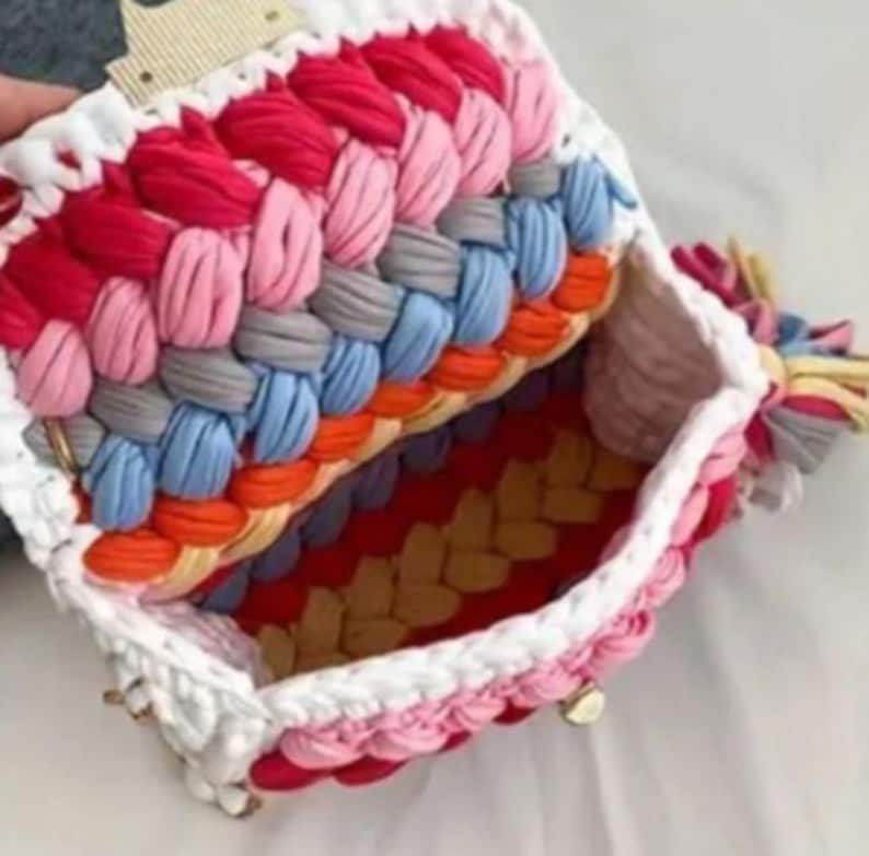 May include: A handmade, open handbag with a colorful, braided design. The bag features a white border and a variety of colors, including red, pink, gray, blue, orange, and yellow. The interior is visible, showcasing a matching color scheme.