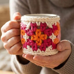 Op de afbeelding: Een handgemaakte mok met een gehaakte hoes. De hoes heeft een bloemenpatroon in roze, oranje en witte tinten. De mok wordt vastgehouden door een persoon die een neutraal gekleurde trui draagt.