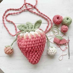 May include: A handmade crochet strawberry-shaped bag in shades of pink and white, with green leaves and a pink cord strap. The bag is accompanied by yarn balls and crochet tools, showcasing the crafting process.