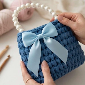 May include: A handmade blue crochet handbag with a pearl beaded handle and a light blue satin bow. The bag is square-shaped and appears to be made of thick yarn. The background includes yarn and crochet hooks.