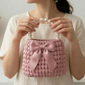 May include: A pink crocheted handbag with a large bow and pearl handles. The bag is a soft pink colour and has a textured appearance. The handles are made of large, round pearls. The bag is held by a person.