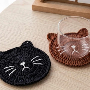 Könnte beinhalten: Zwei handgefertigte Untersetzer in Katzenform. Einer ist schwarz, der andere braun. Beide haben weiße gestickte Details für das Gesicht. Eine transparente Glas-Tasse steht auf dem braunen Untersetzer. Die Untersetzer liegen auf einer hellen Holzoberfläche.
